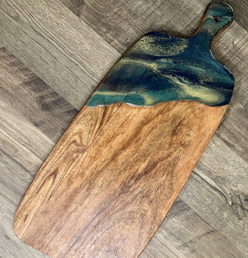 Rectangular acacia wood cutting board with handle with hand poured resin in dark green and blue hues and a ribbon of gold.